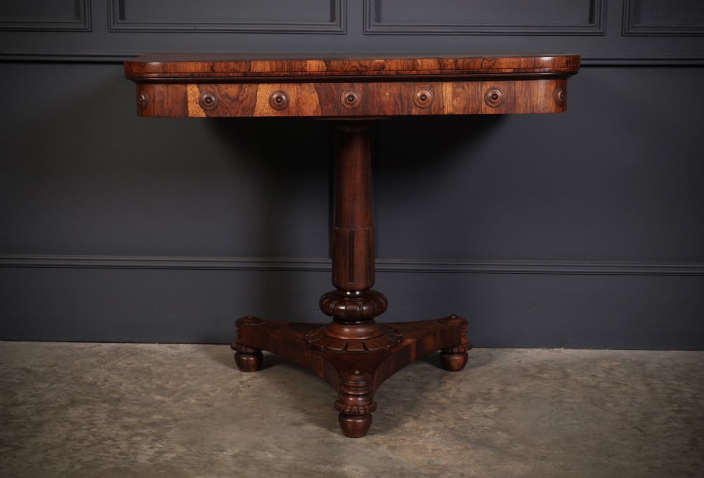 An antique Regency rosewood card table featuring a hinged rectangular top, rosette-decorated frieze, turned column, and triform pedestal base. View VII