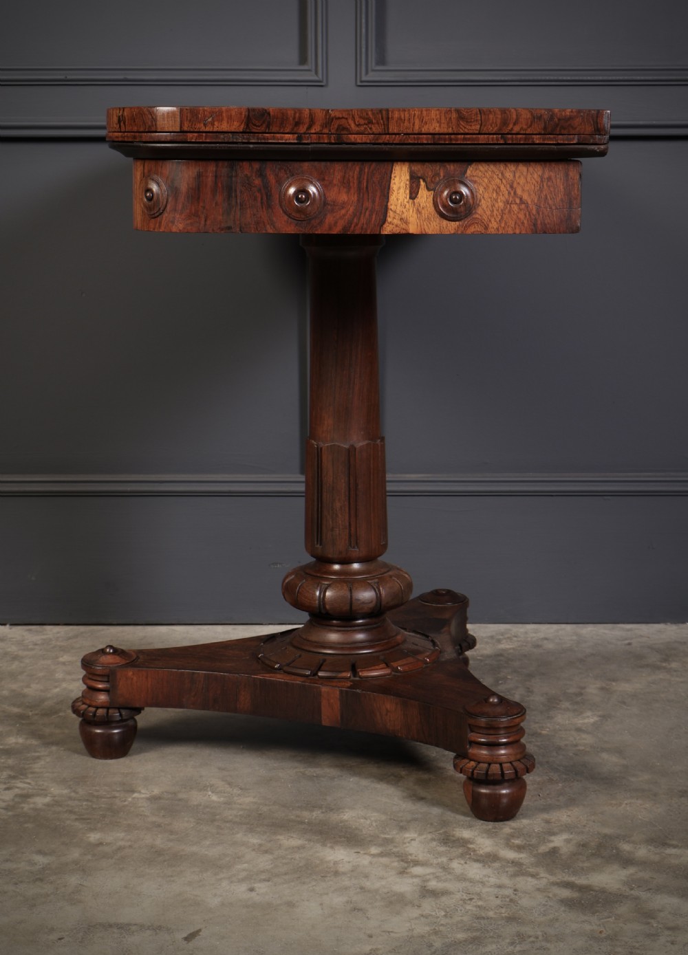 An antique Regency rosewood card table featuring a hinged rectangular top, rosette-decorated frieze, turned column, and triform pedestal base. View XI