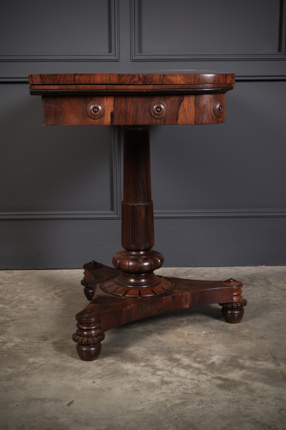 An antique Regency rosewood card table featuring a hinged rectangular top, rosette-decorated frieze, turned column, and triform pedestal base. View XIII