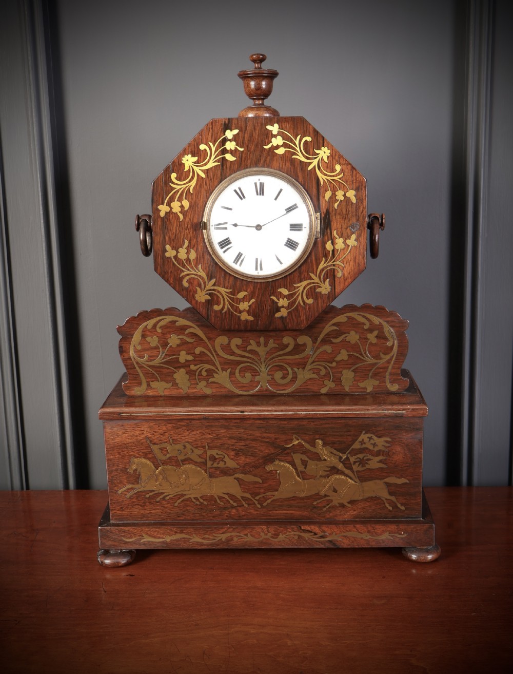 A Regency dark rosewood mantel clock with intricate brass inlay, an octagonal dial surround, and an equestrian brass scene on the base plinth.