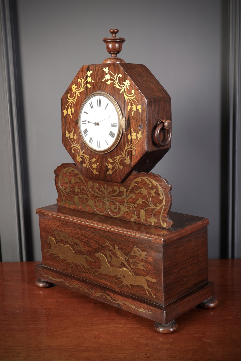 A Regency dark rosewood mantel clock with intricate brass inlay, an octagonal dial surround, and an equestrian brass scene on the base plinth. View VI
