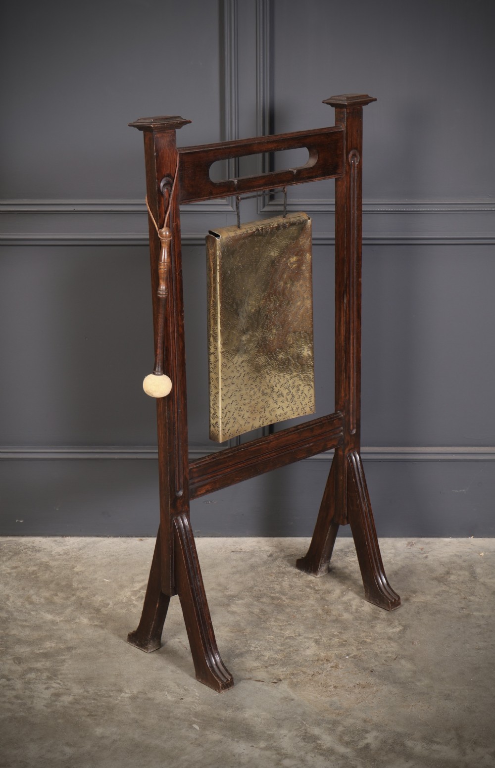 A freestanding dark oak frame housing a suspended rectangular hammered brass dinner gong complete with a wooden striker.