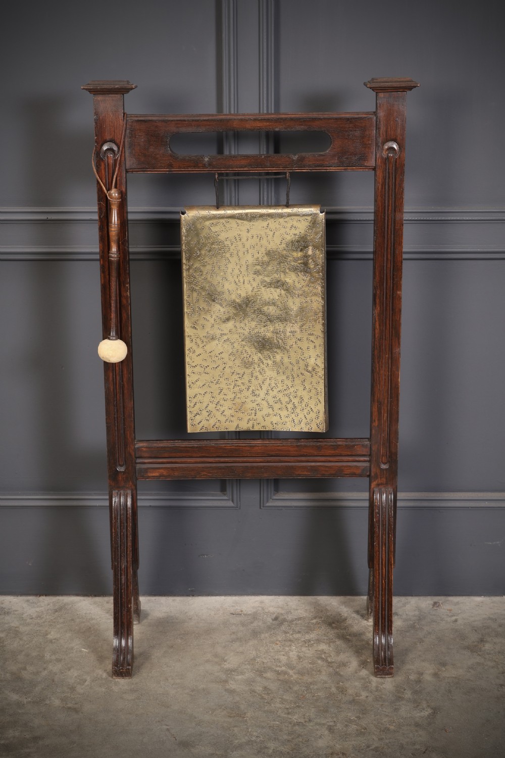 A freestanding dark oak frame housing a suspended rectangular hammered brass dinner gong complete with a wooden striker. View II