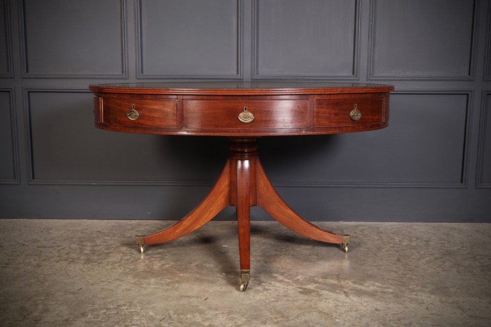 A Georgian mahogany oval drum table with a green leather inset top, frieze drawers with circular brass pulls, and a central pedestal resting on four splayed legs with brass castors. View II