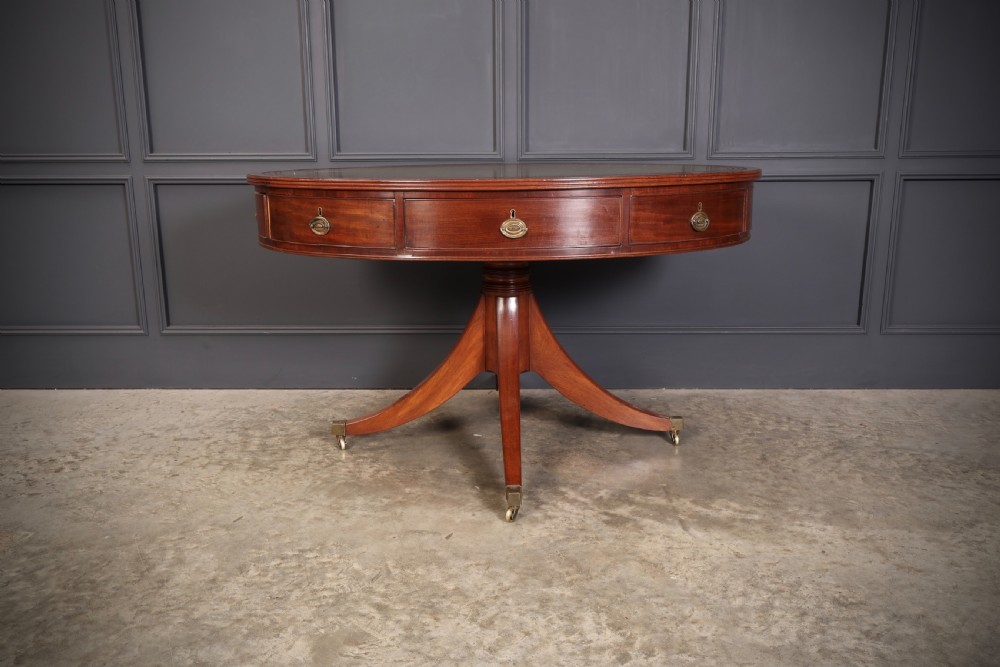A Georgian mahogany oval drum table with a green leather inset top, frieze drawers with circular brass pulls, and a central pedestal resting on four splayed legs with brass castors. View V