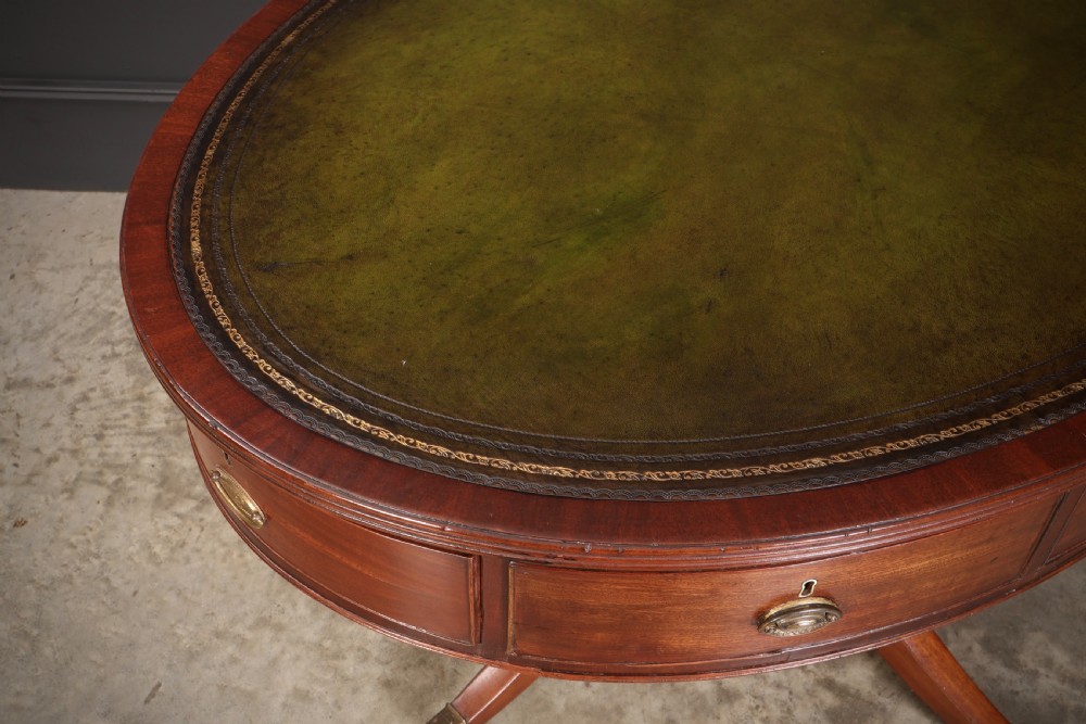 A Georgian mahogany oval drum table with a green leather inset top, frieze drawers with circular brass pulls, and a central pedestal resting on four splayed legs with brass castors. View VII