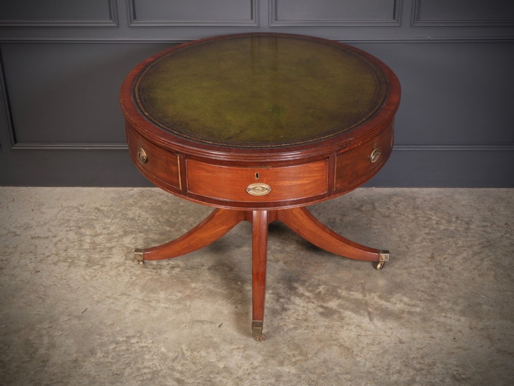 A Georgian mahogany oval drum table with a green leather inset top, frieze drawers with circular brass pulls, and a central pedestal resting on four splayed legs with brass castors. View IX