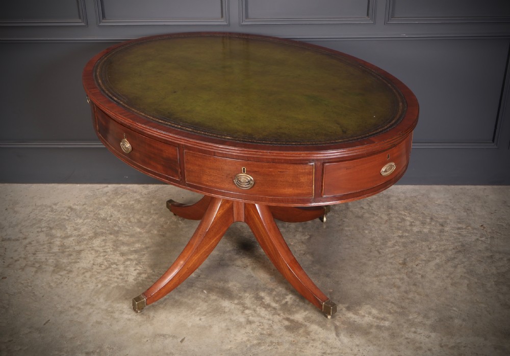 A Georgian mahogany oval drum table with a green leather inset top, frieze drawers with circular brass pulls, and a central pedestal resting on four splayed legs with brass castors. View X