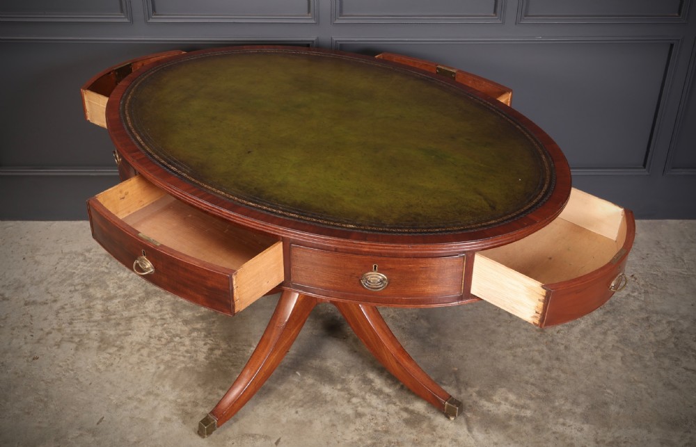 A Georgian mahogany oval drum table with a green leather inset top, frieze drawers with circular brass pulls, and a central pedestal resting on four splayed legs with brass castors. View XI