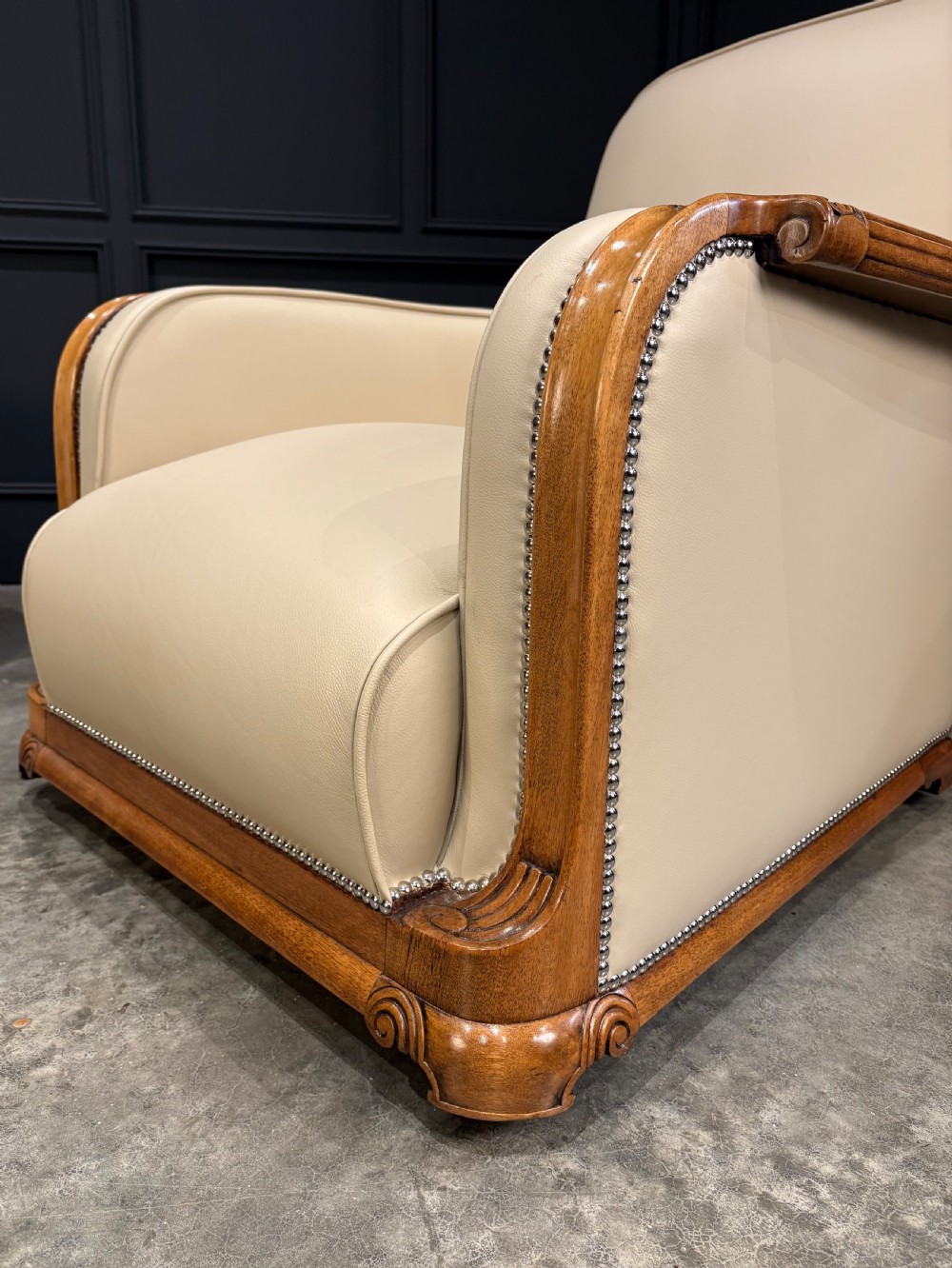 An Art Deco three-piece suite featuring a three-seater sofa and two armchairs upholstered in cream leather with polished walnut trim and stud details. View IV