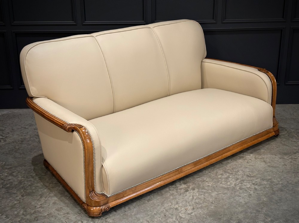 An Art Deco three-piece suite featuring a three-seater sofa and two armchairs upholstered in cream leather with polished walnut trim and stud details. View XV