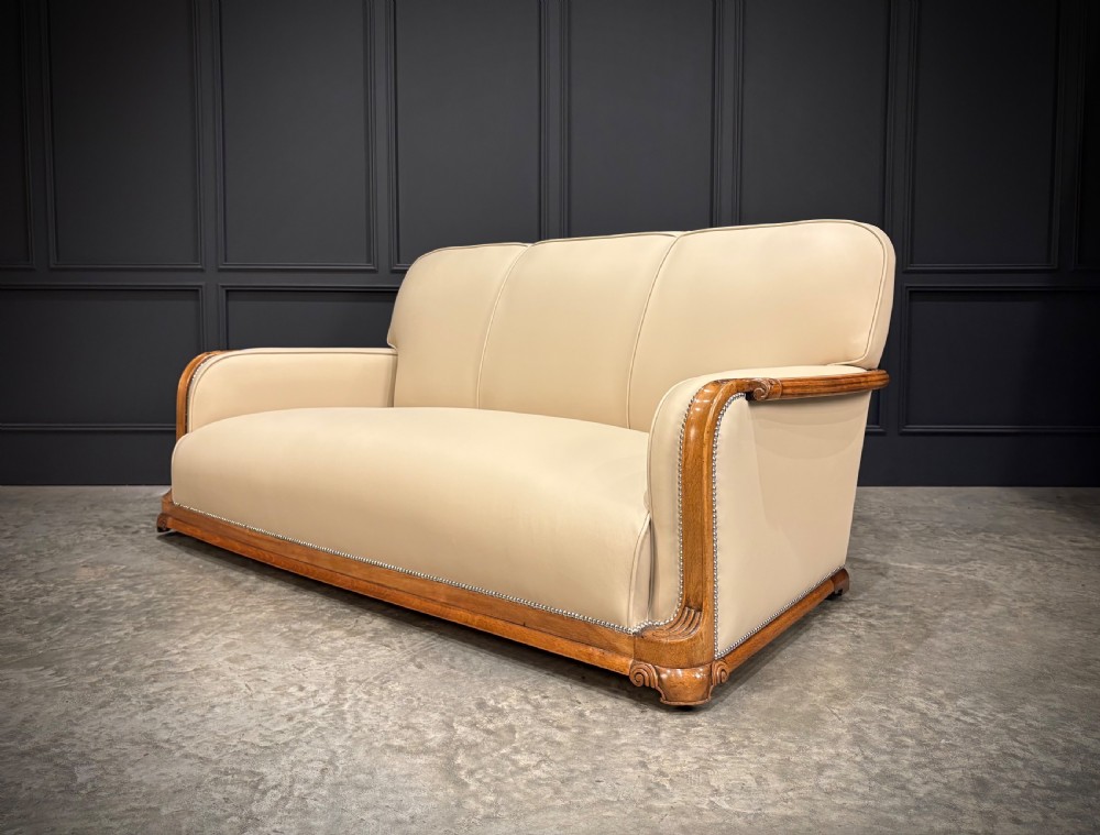 An Art Deco three-piece suite featuring a three-seater sofa and two armchairs upholstered in cream leather with polished walnut trim and stud details. View XVIII