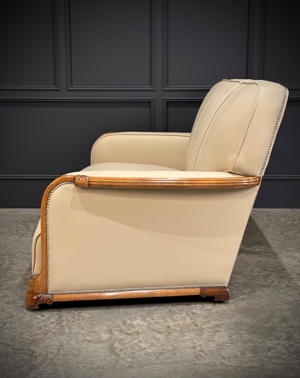 An Art Deco three-piece suite featuring a three-seater sofa and two armchairs upholstered in cream leather with polished walnut trim and stud details. View XX