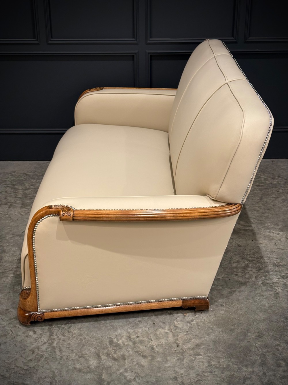 An Art Deco three-piece suite featuring a three-seater sofa and two armchairs upholstered in cream leather with polished walnut trim and stud details. View XXI