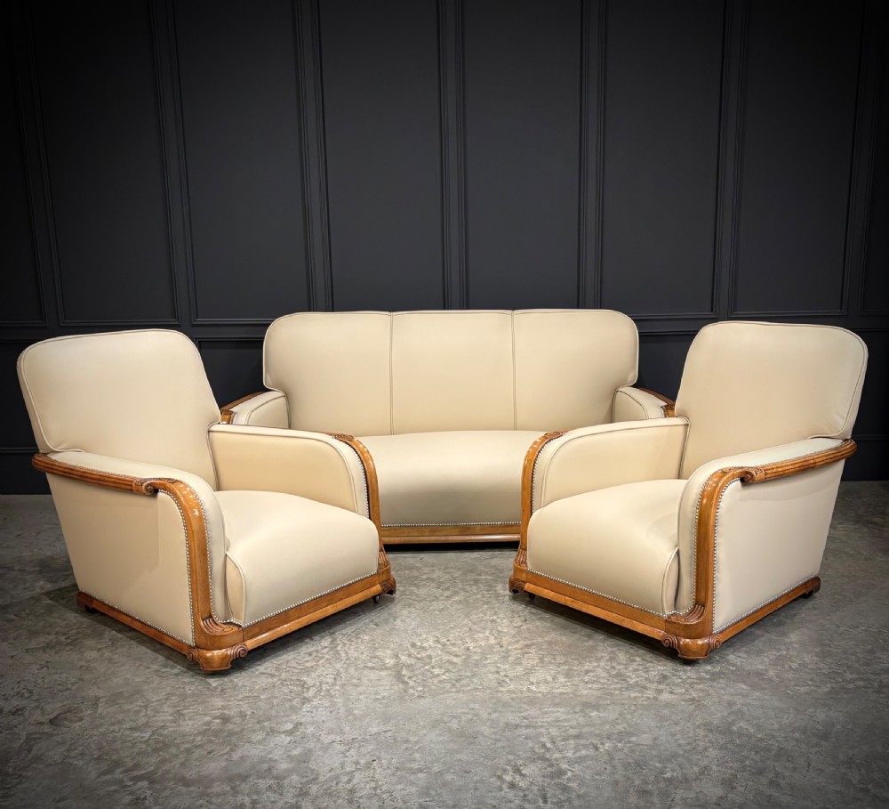 An Art Deco three-piece suite featuring a three-seater sofa and two armchairs upholstered in cream leather with polished walnut trim and stud details.