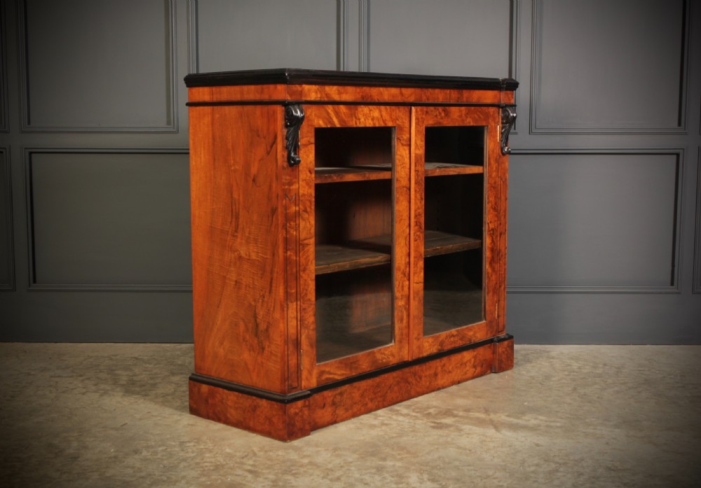 A low Victorian bookcase constructed from figured walnut wood featuring two glazed glass doors, black carved corbels, and internal timber shelves.