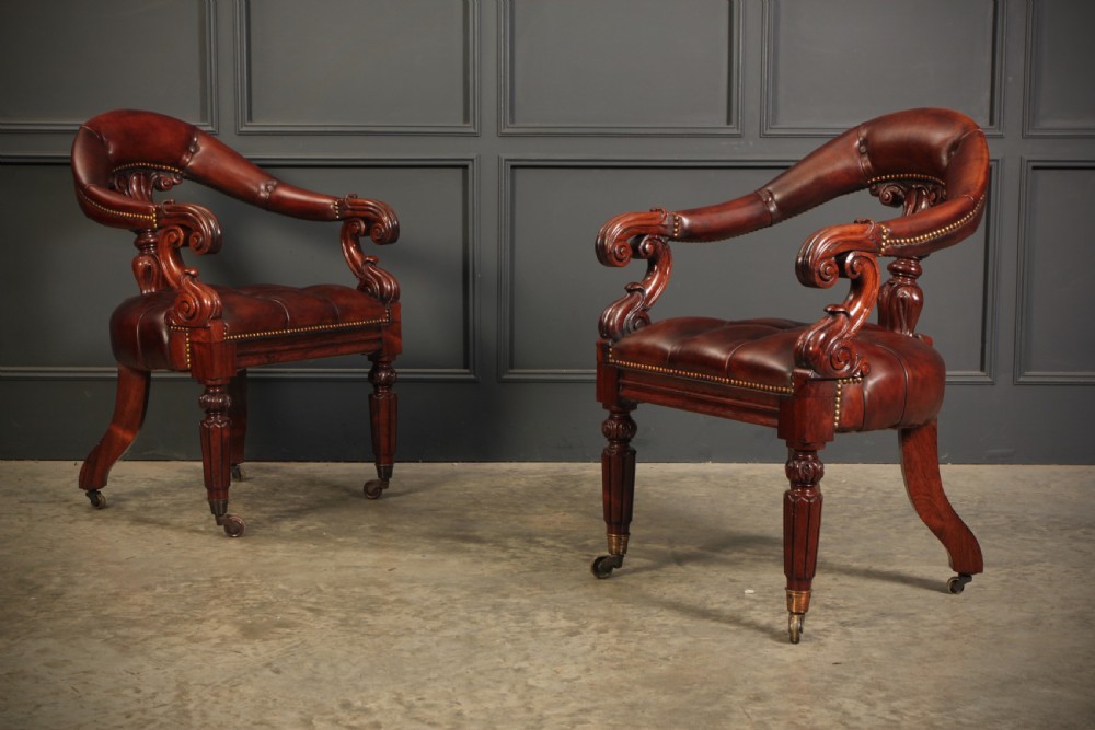 A pair of William IV rosewood desk chairs featuring deep buttoned reddish-brown leather upholstery, carved scroll arms, turned front legs with brass castors, and splayed rear legs.