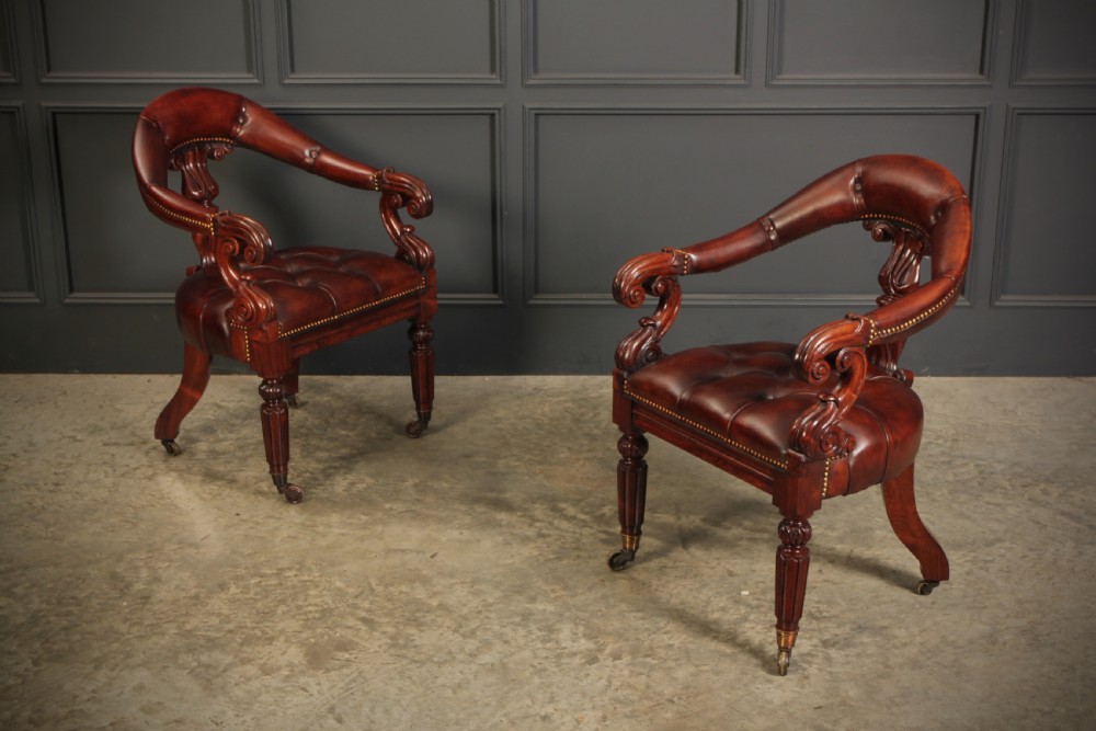A pair of William IV rosewood desk chairs featuring deep buttoned reddish-brown leather upholstery, carved scroll arms, turned front legs with brass castors, and splayed rear legs. View II