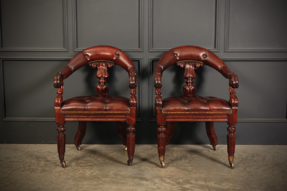 A pair of William IV rosewood desk chairs featuring deep buttoned reddish-brown leather upholstery, carved scroll arms, turned front legs with brass castors, and splayed rear legs. View III