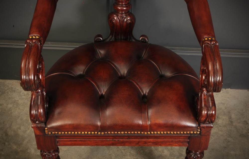 A pair of William IV rosewood desk chairs featuring deep buttoned reddish-brown leather upholstery, carved scroll arms, turned front legs with brass castors, and splayed rear legs. View VI