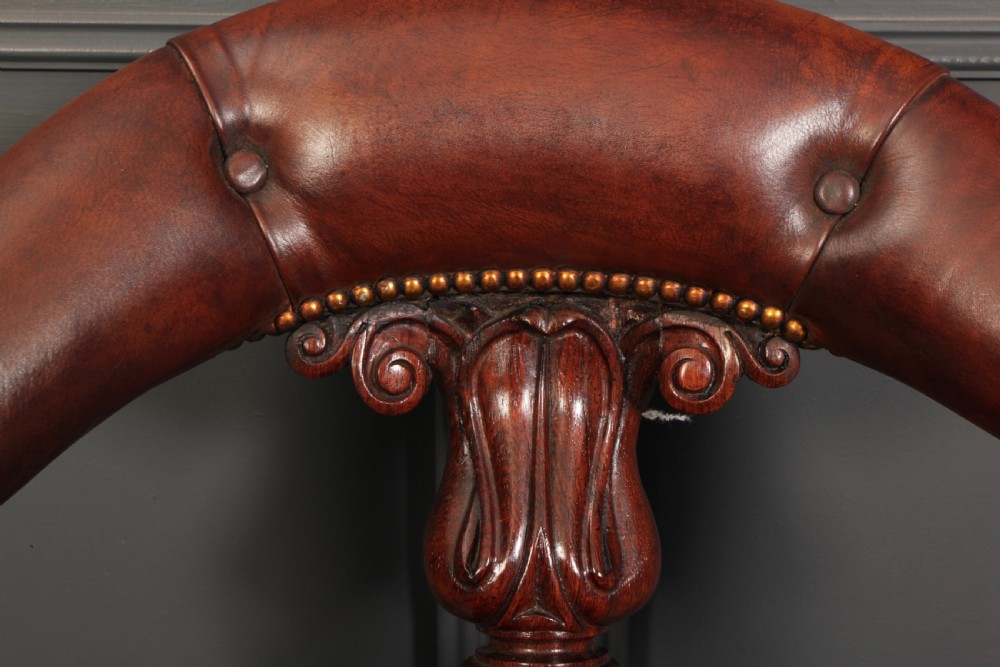 A pair of William IV rosewood desk chairs featuring deep buttoned reddish-brown leather upholstery, carved scroll arms, turned front legs with brass castors, and splayed rear legs. View VII