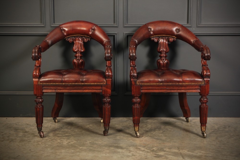 A pair of William IV rosewood desk chairs featuring deep buttoned reddish-brown leather upholstery, carved scroll arms, turned front legs with brass castors, and splayed rear legs. View XIII