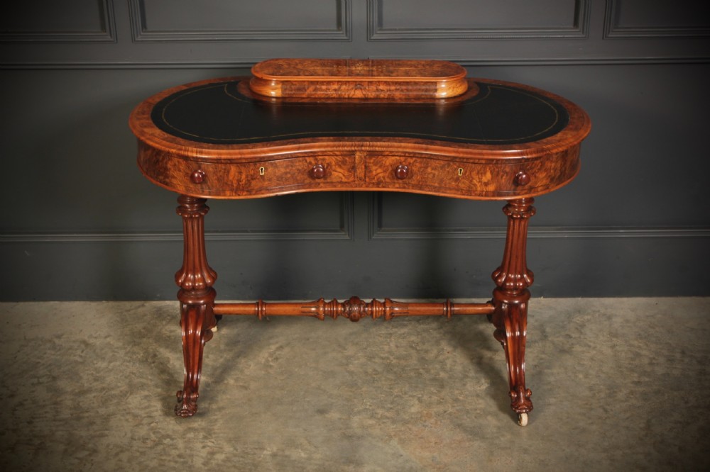 A Victorian burr walnut kidney-shaped writing table featuring a black leather insert, inlaid marquetry, raised caddy, frieze drawers, and carved legs on castors.