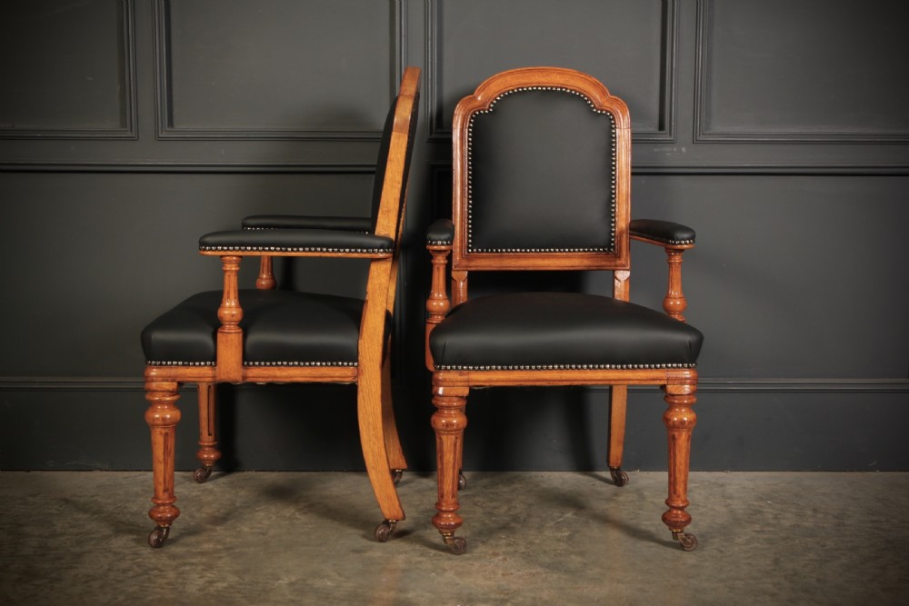 A pair of Victorian oak desk armchairs upholstered in black leather with brass stud trimming and turned legs on castors. View V