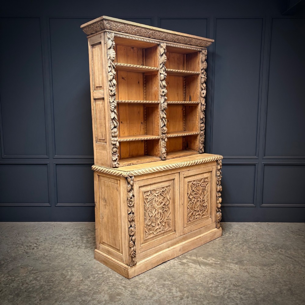 A large two-tier bleached oak open bookcase featuring heavily carved floral pilasters, open adjustable shelves, and a lower cabinet with relief-carved doors.