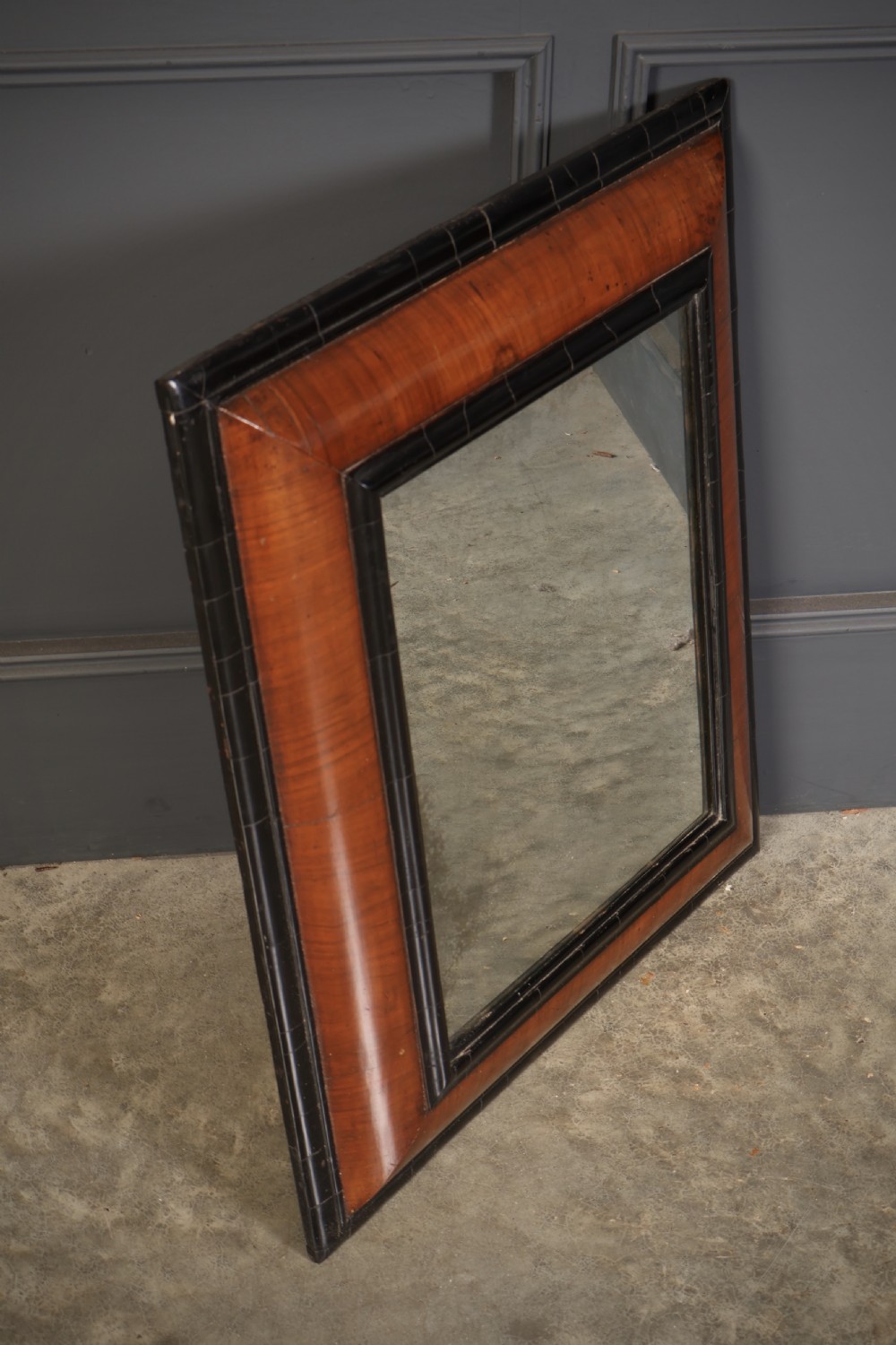 A rectangular antique cushion frame mirror featuring a warm walnut veneer section enclosed by dark ebonised mouldings around an aged glass plate. View II