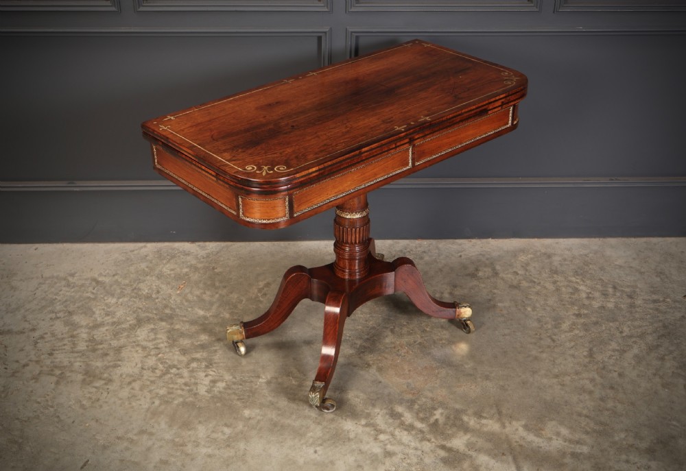 An antique Regency rosewood fold-over card table featuring brass inlay details, a turned pedestal stem, and four swept legs with brass castors.