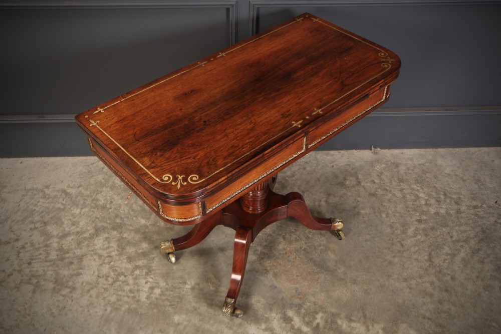 An antique Regency rosewood fold-over card table featuring brass inlay details, a turned pedestal stem, and four swept legs with brass castors. View II