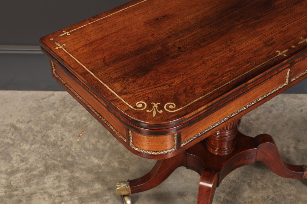 An antique Regency rosewood fold-over card table featuring brass inlay details, a turned pedestal stem, and four swept legs with brass castors. View III
