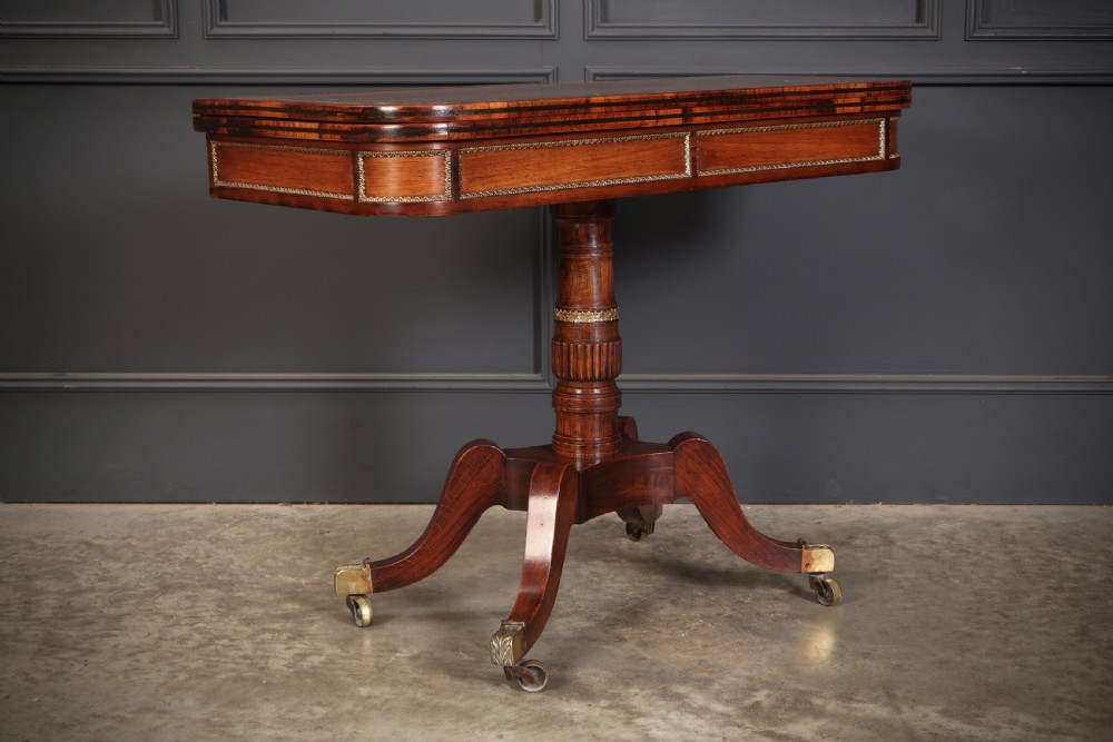 An antique Regency rosewood fold-over card table featuring brass inlay details, a turned pedestal stem, and four swept legs with brass castors. View V