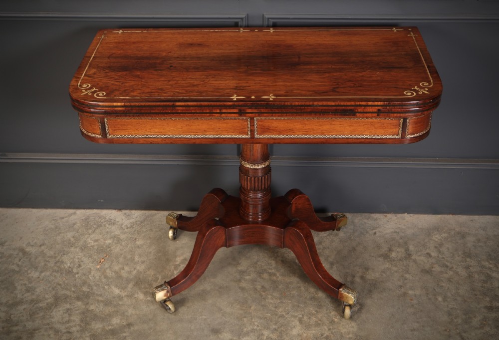 An antique Regency rosewood fold-over card table featuring brass inlay details, a turned pedestal stem, and four swept legs with brass castors. View VI