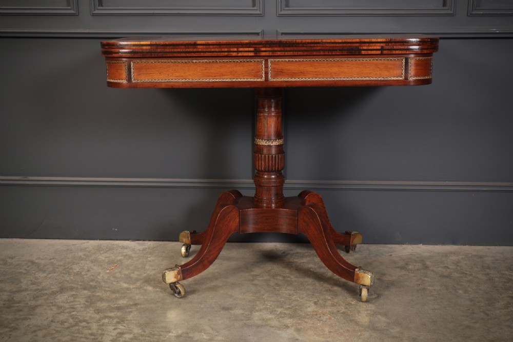 An antique Regency rosewood fold-over card table featuring brass inlay details, a turned pedestal stem, and four swept legs with brass castors. View VII
