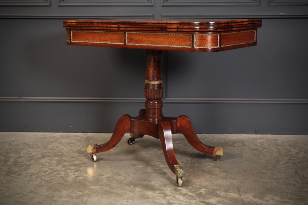 An antique Regency rosewood fold-over card table featuring brass inlay details, a turned pedestal stem, and four swept legs with brass castors. View X