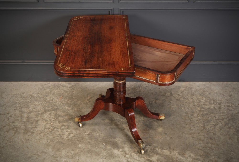 An antique Regency rosewood fold-over card table featuring brass inlay details, a turned pedestal stem, and four swept legs with brass castors. View XII