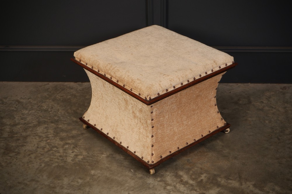 A square Regency box ottoman upholstered in textured beige fabric with dark mahogany trim, brass stud detailing, and small wheel castors.
