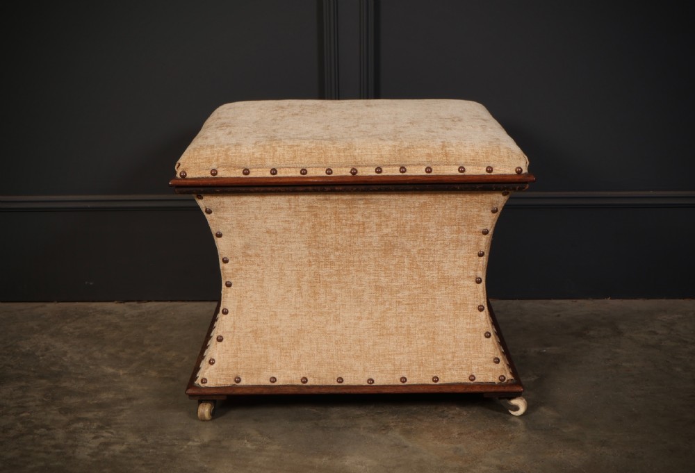 A square Regency box ottoman upholstered in textured beige fabric with dark mahogany trim, brass stud detailing, and small wheel castors. View V