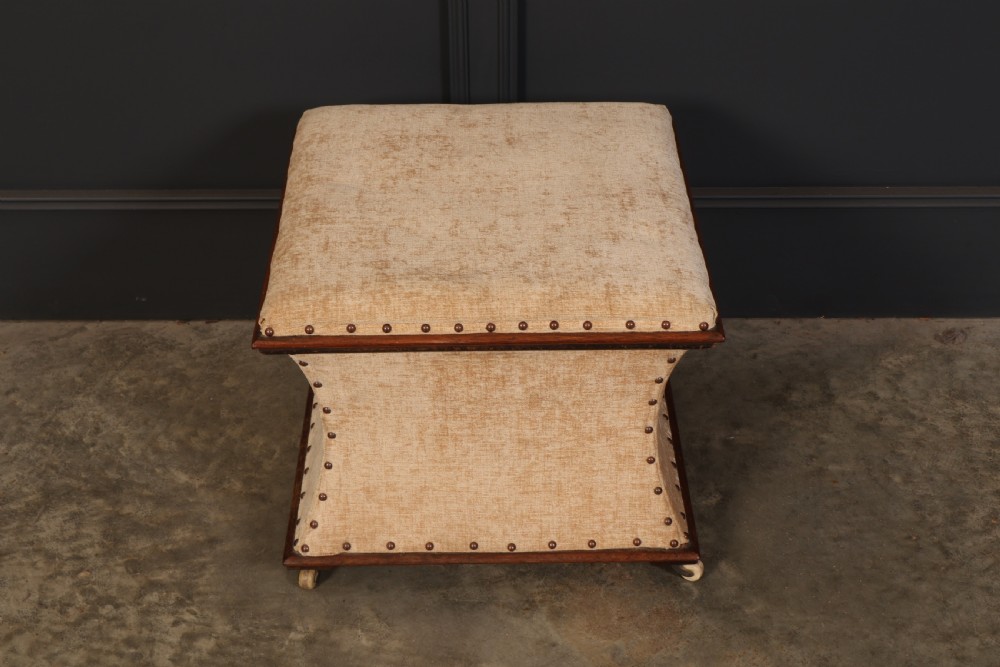 A square Regency box ottoman upholstered in textured beige fabric with dark mahogany trim, brass stud detailing, and small wheel castors. View VI