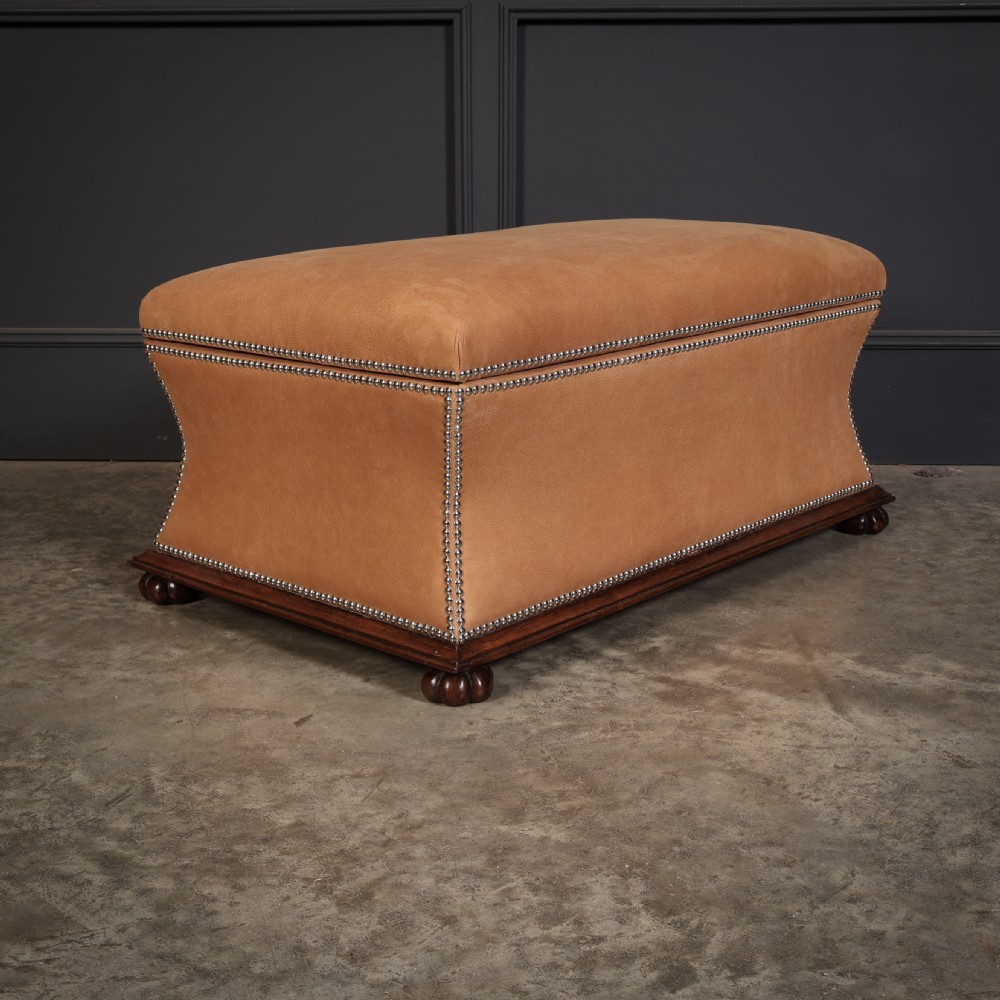 A rectangular tan leather upholstered box ottoman featuring brass stud trim and concave sides on a dark rosewood plinth base with turned feet. View VII