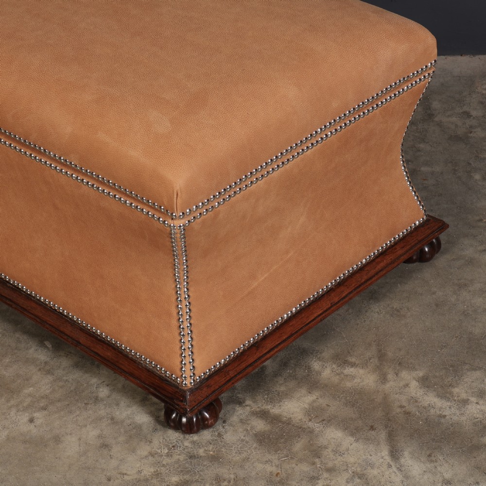 A rectangular tan leather upholstered box ottoman featuring brass stud trim and concave sides on a dark rosewood plinth base with turned feet. View XI