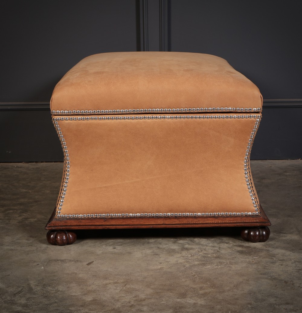 A rectangular tan leather upholstered box ottoman featuring brass stud trim and concave sides on a dark rosewood plinth base with turned feet. View XII