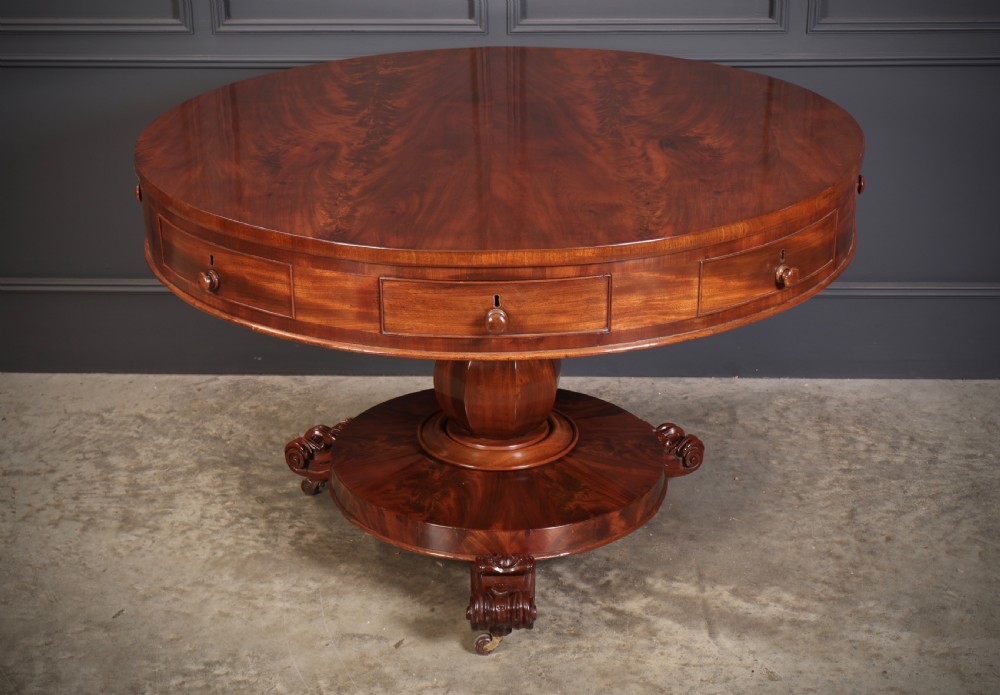 A circular William IV mahogany drum table featuring a flame mahogany top, frieze drawers with wooden knobs, a thick central column, and a circular plinth base with carved scroll feet.