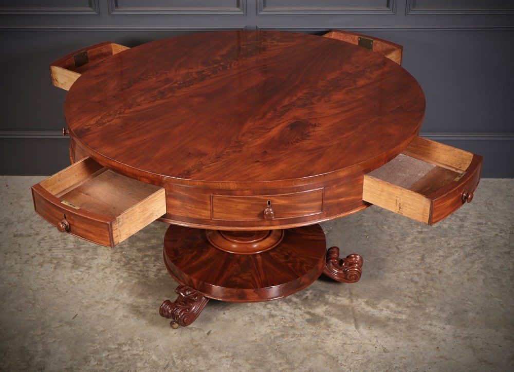 A circular William IV mahogany drum table featuring a flame mahogany top, frieze drawers with wooden knobs, a thick central column, and a circular plinth base with carved scroll feet. View III