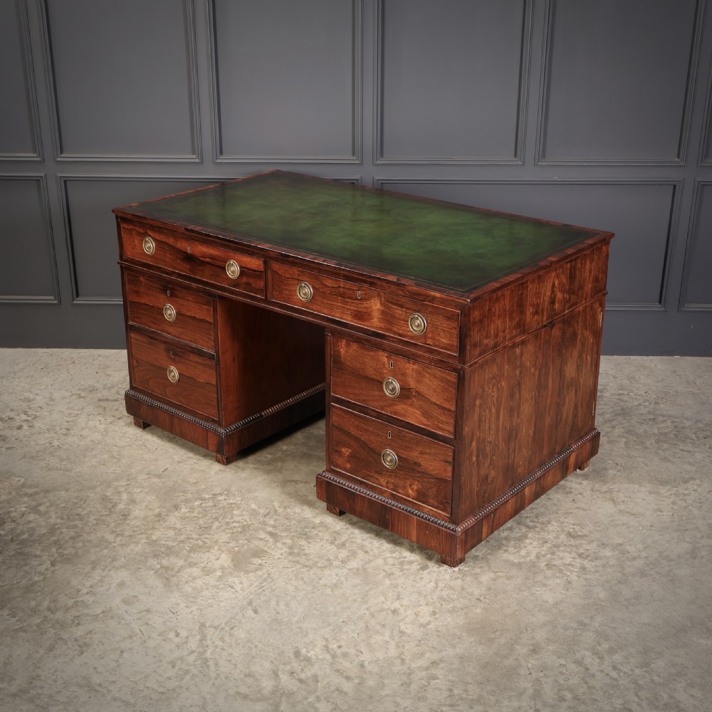 A William IV rosewood pedestal desk featuring a green leather writing surface, multiple drawers with brass ring handles, and decorative moulding around the lower base.