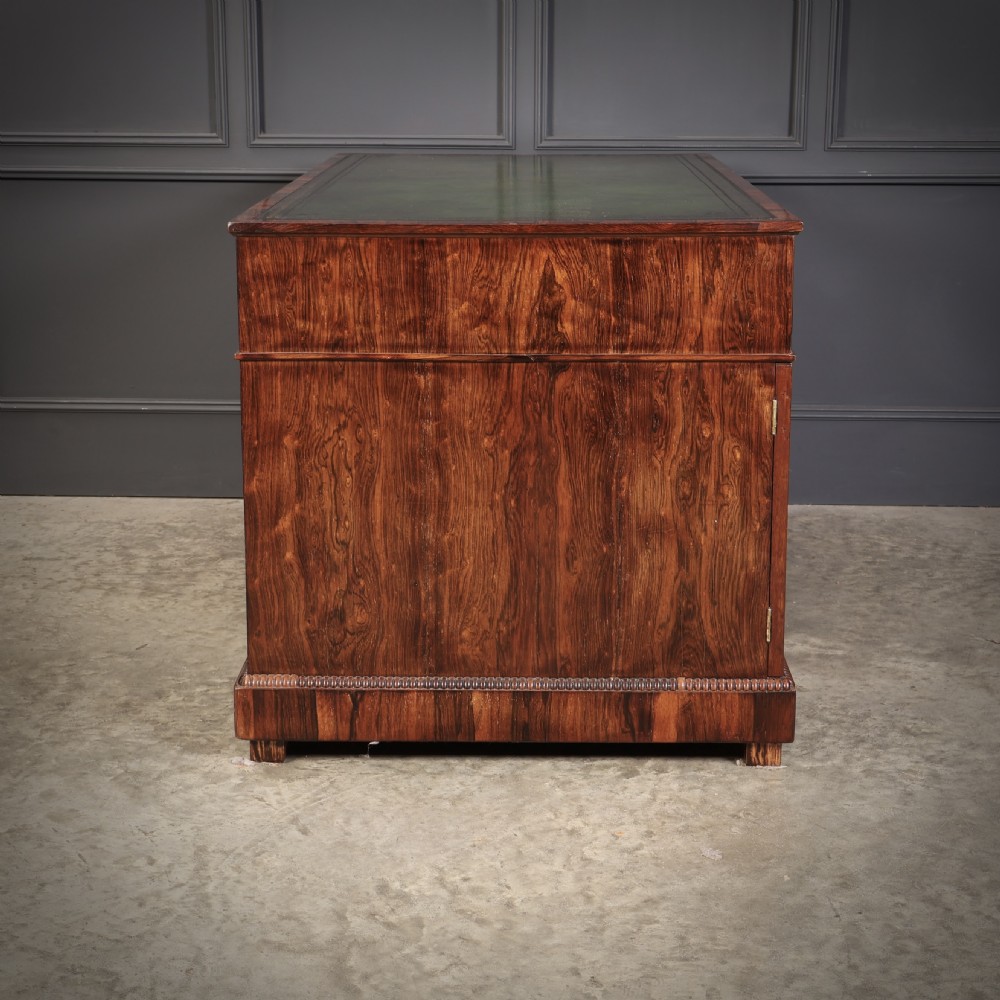 A William IV rosewood pedestal desk featuring a green leather writing surface, multiple drawers with brass ring handles, and decorative moulding around the lower base. View X