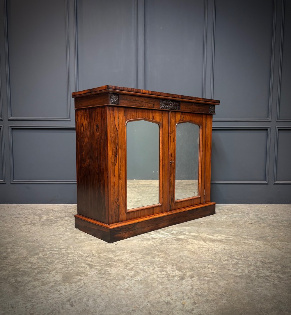 A William IV rosewood side cabinet with twin arch-topped mirrored doors, carved foliage details along the frieze, and a sturdy plinth base.