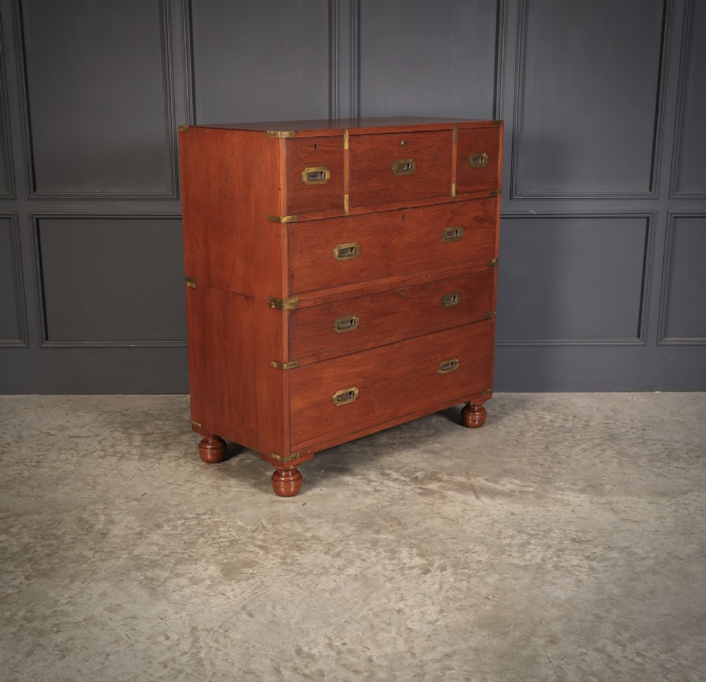 A Victorian teak military campaign secretaire chest featuring an open fall-front desk compartment, flush brass handles, brass corner brackets, and turned feet against a dark grey panelled wall. View VII